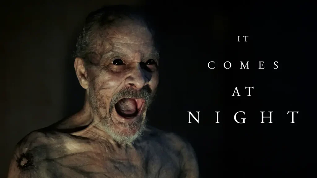 Dark promotional poster for It Comes at Night (2017), featuring a frightening elderly man with black eyes and an open mouth against a shadowy background.